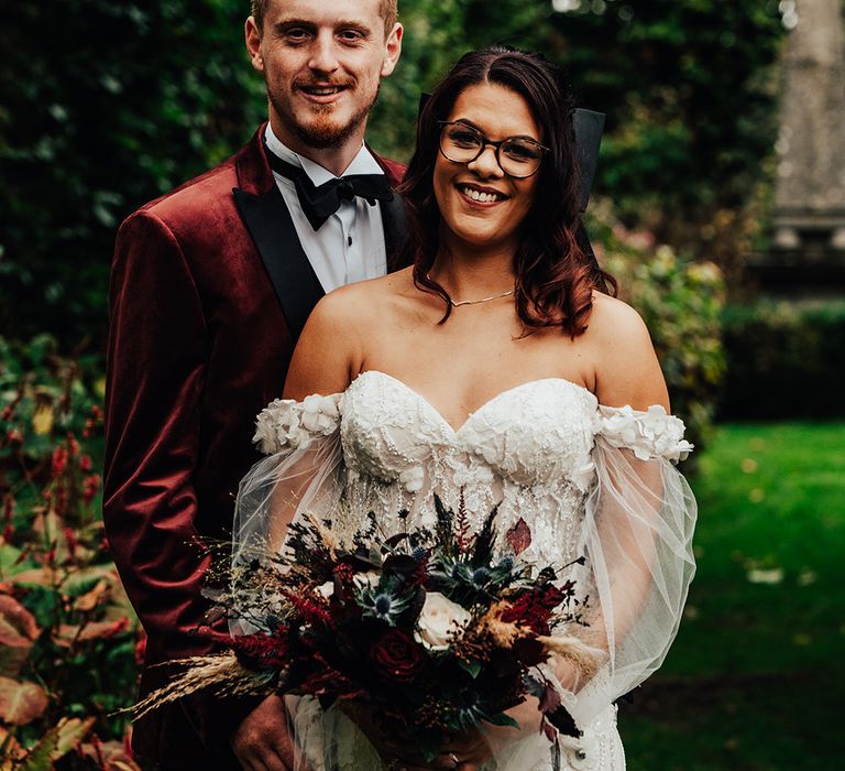 Groom in burgundy velvet suit with black tie standing with bride in embellished wedding dress with black bow hair accessory 