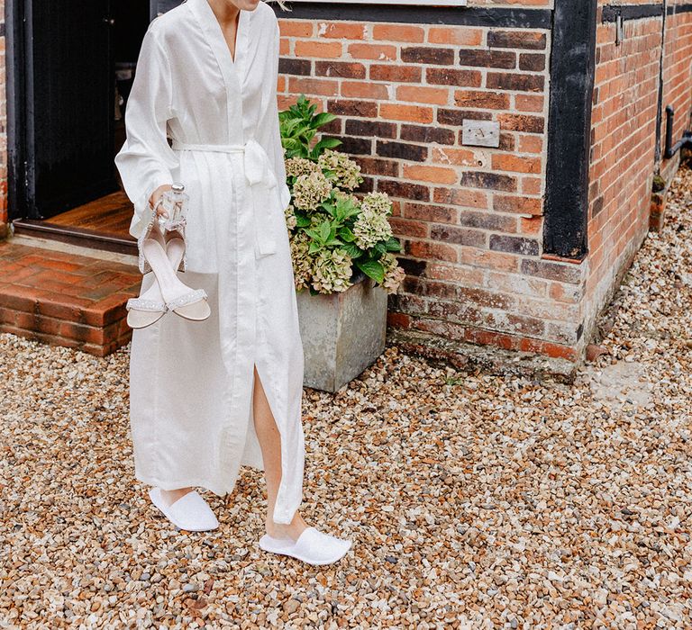 Bride in long white satin robe wearing white slippers carrying her sparkly wedding shoes and Jo Malone wedding scent 