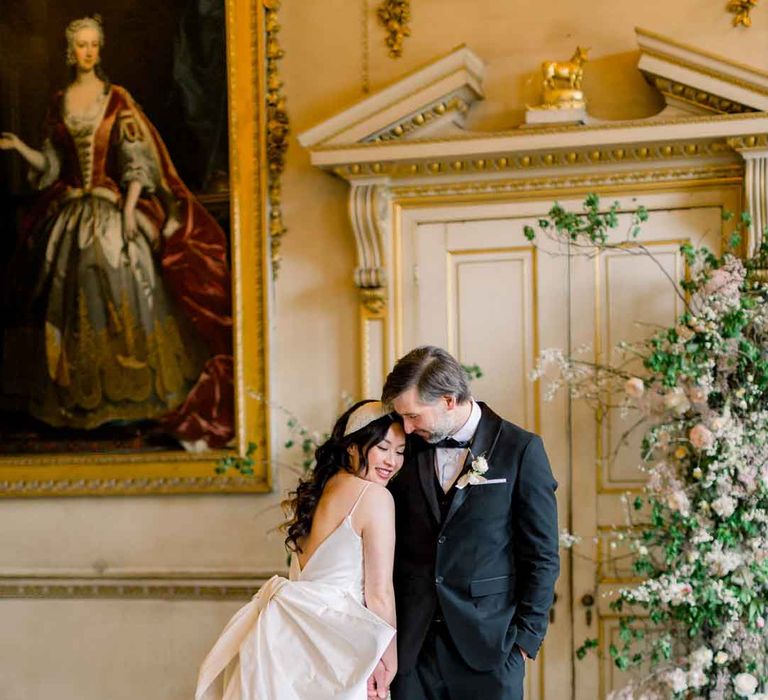 Bride in sleeveless satin wedding dress with large accent bow on the back and groom in classic black tuxedo, black bowtie and boutonniere    