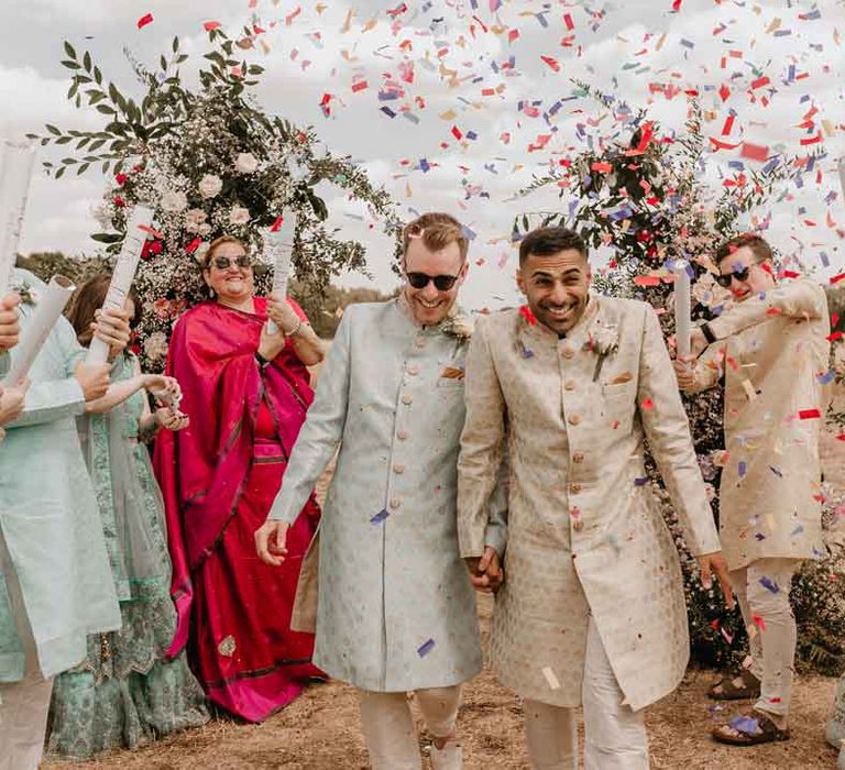 Groom in mint green Indian Sherwani with white garden rose and foliage boutonniere and grooms sunglasses holding hands with groom in cream and gold Indian sherwani and white garden rose boutonniere doing colourful wedding confetti exit at Gujarati wedding ceremony at High Billinghurst Farm