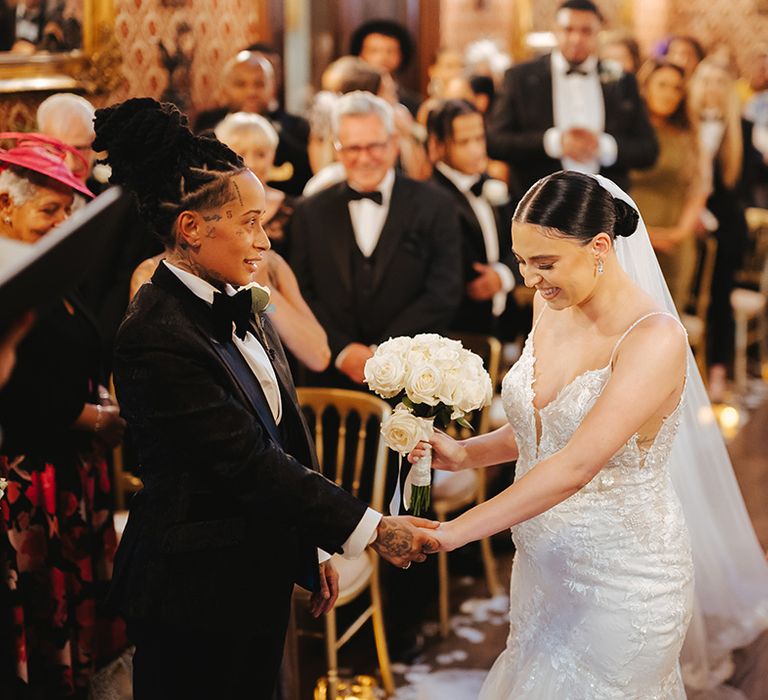 Bride holding white rose bridal bouquet wearing sleeveless v neck Enzoani wedding dress with tulle finish and church length veil holding hands with bride in black snakeskin print tuxedo with black bowtie and white rose boutonniere at the alter of Grittleton House