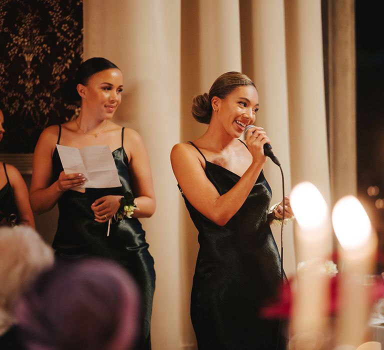 Bridesmaids in strappy satin midi length black bridesmaid dresses doing wedding speeches at Grittleton House wedding venue 