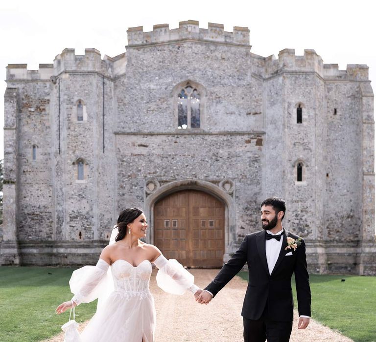 Bride in ethereal off the shoulder wedding dress with detachable puff tulle sleeves and embroidered rose 3d appliqué details walking hand in hand with groom in classic black tuxedo with bowtie and garden rose and eucalyptus boutonniere outside Pentney Abbey wedding venue 