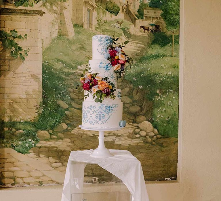 Four tiered toile wedding cake in blue and white on white cake stand on clear glass stand with white wedding drapes and bright blue glass bottles with dried flowers