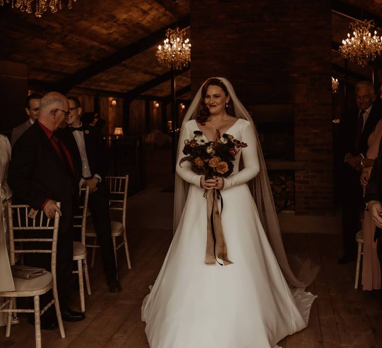 Bride in long sleeve deep v wedding dress and church length veil walking down the aisle at Hidden River Cabins holding white, dusky pink and burnt red dried flower, rose and peony bridal bouquet 