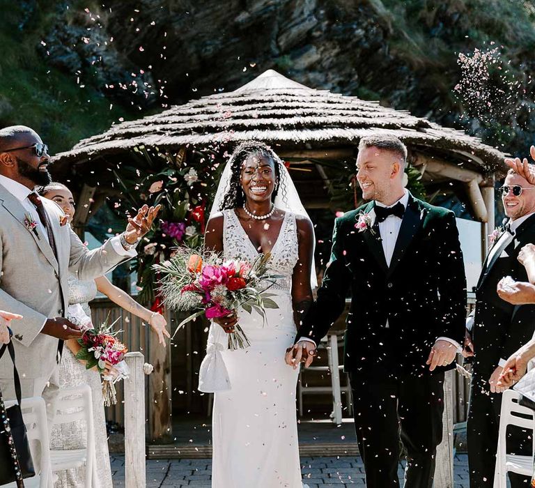 Bride in v neck sleeveless lace Pronovias wedding dress, cathedral length veil and silver sparkly bridal jewellery doing confetti exit with groom in bottle green velvet grooms blazer, colourful tropical boutonniere and black suit trousers 