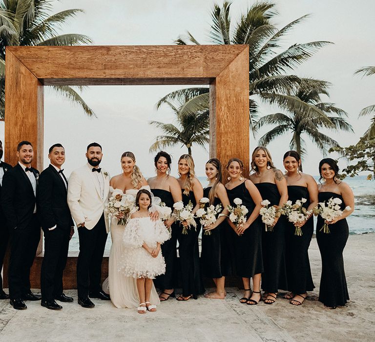 wedding party photography at Blue Venado beach wedding in front of a wooden Frame with black bridesmaid dresses and tuxedos 