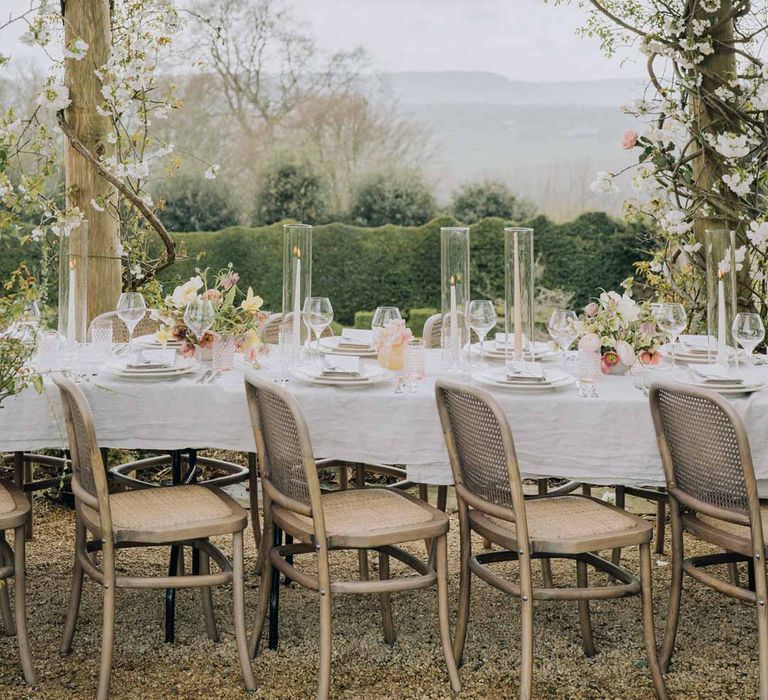 Outdoor al fresco wedding tablescape with blush pink tapered candles in glass candlestick covers on spring inspired wedding tablescape with white ruffled wedding table cloth, white peony, pink garden rose, foliage and blush florals wedding table centrepieces and simple crockery at Euridge Manor wedding