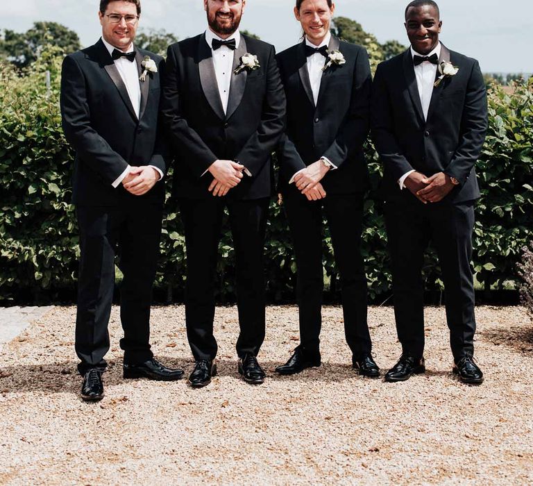 Grooms party wearing matching classic black three piece grooms tuxedos with white rose boutonnieres and black bowties 