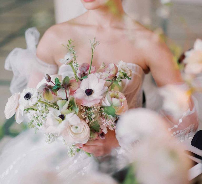 Bride in sweetheart neckline tulle mesh bridal gown with detachable puff tulle sleeves and delicate beading holding bridal bouquet with white poppies, light pink Persian buttercups, baby's-breath and foliage 