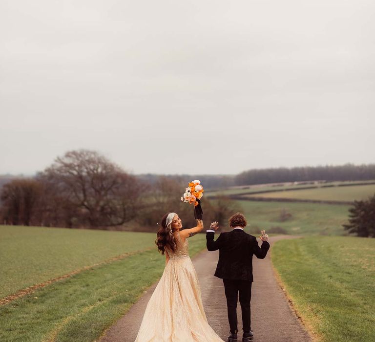 Bride in sleeveless deep v-neck sparkly gold wedding dress holding up orange garden rose, white poppy, light pink carnation and foliage bridal bouquet wrapped in sparkly gold ribbon with bride in dark bridal suit at Kingscote Barn Tetbury 