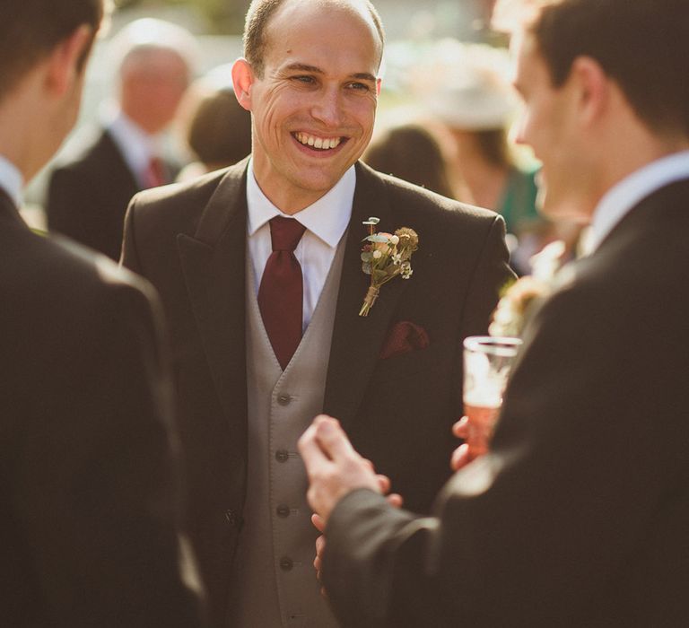 Groom in dark suit with light grey waistcoat and red tie with floral buttonhole socialising with guests 