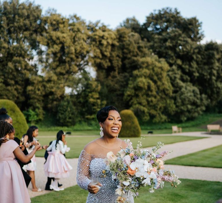 Bride in illusion v-neck sheer off shoulder long sleeve pearl embellished wedding gown holding large pastel and foliage mixed bridal bouquet on the grounds of Syon Park