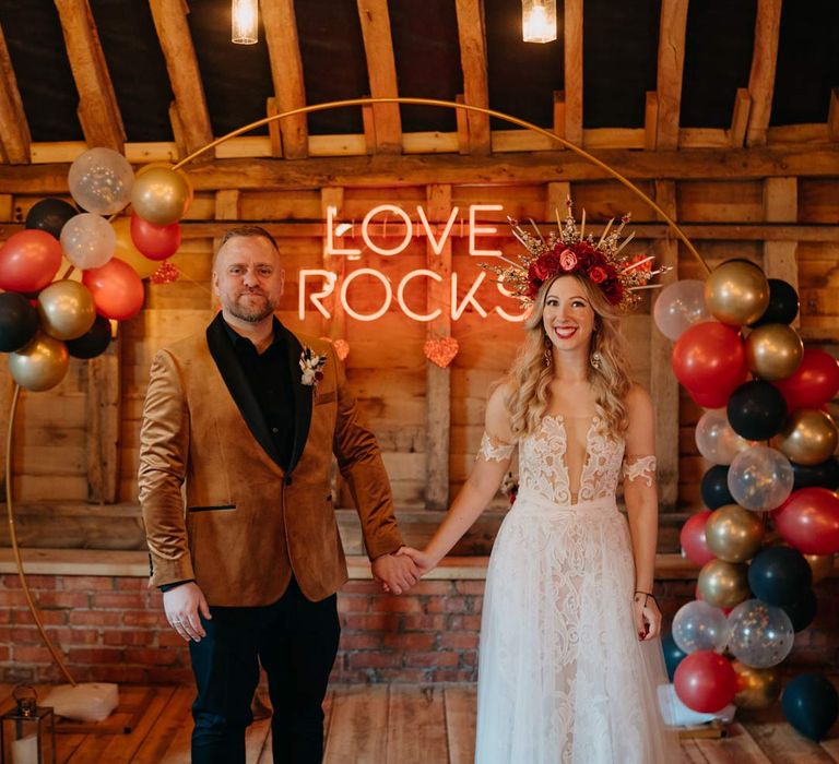 Groom in crushed velvet burnt orange grooms blazer and dried flower boutonniere holding hands with bride in off shoulder lace wedding dress with corset style top and red rose and gold halo bridal crown 