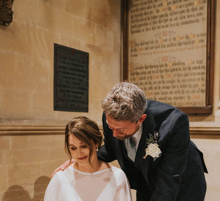 Bride sits to sig the wedding register with the groom leaning over her to look 