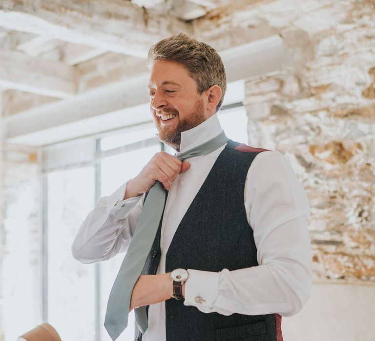 Groom does up his pale green wedding tie as he gets ready for the wedding day 