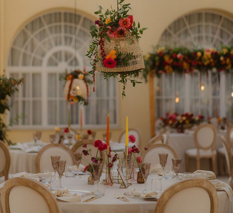 orange and yellow wedding table decorations with wicker lampshade, coloured goblets, taper candles and flower stems in vessels. 