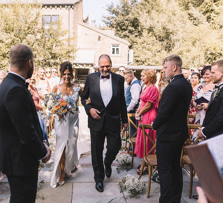 Father of the bride in black tuxedo walks the bride down the aisle at outdoor wedding 