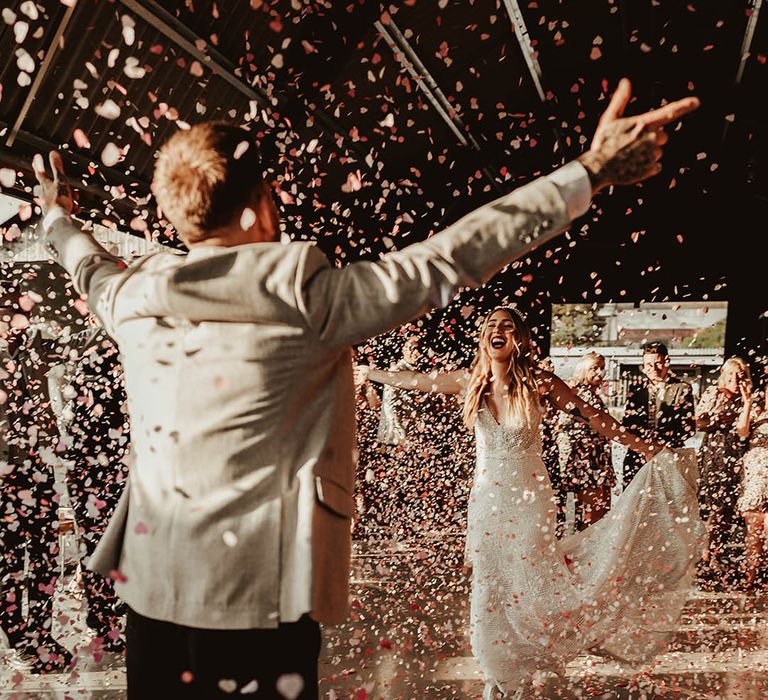 Bride and groom walk towards each other with open arms as pink and red confetti falls around them 