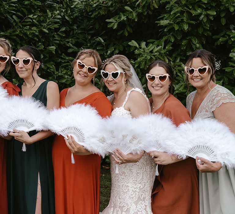 The bride and bridesmaids wear mismatched bridesmaid dresses with feather fans and sunglasses 