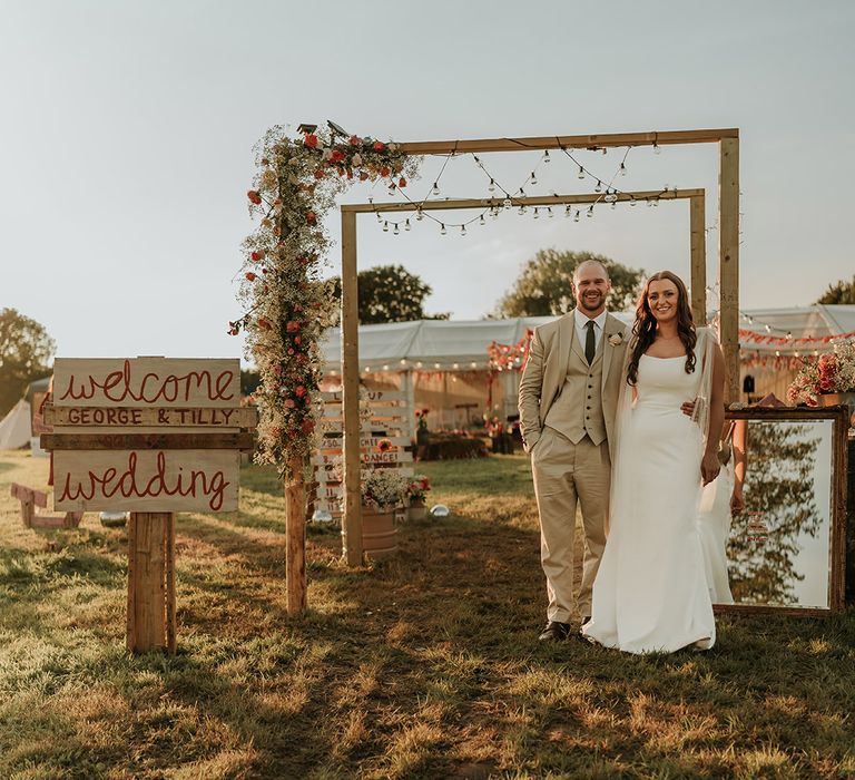 At home summer wedfest wedding with DIY wooden signage as the bride and groom stand under a wooden arch 