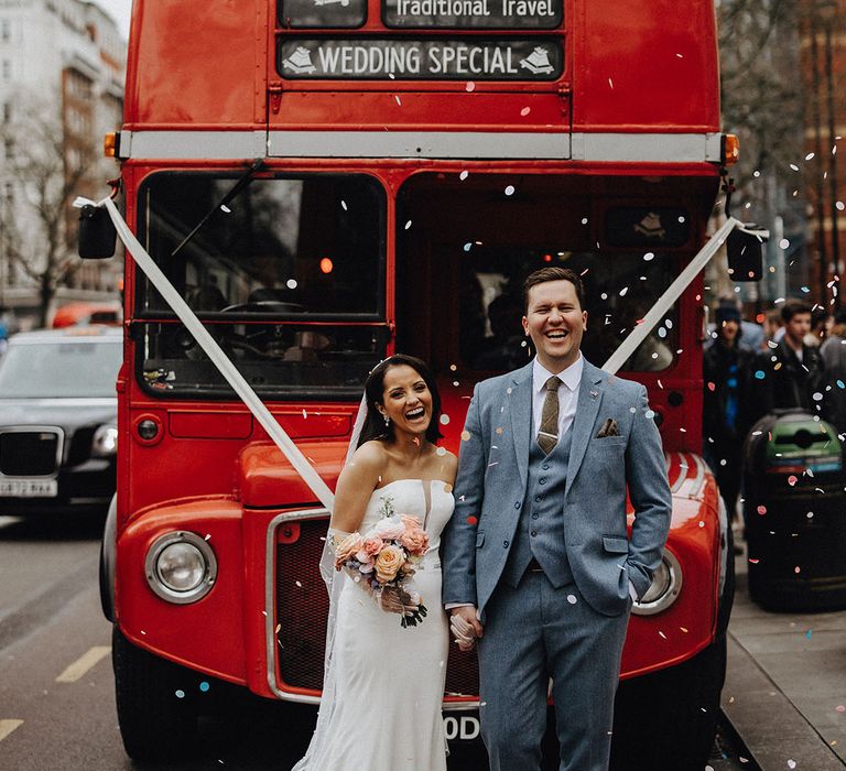 Bride and groom stand in front of their iconic red double decker bus wedding transport with white ribbon decoration 