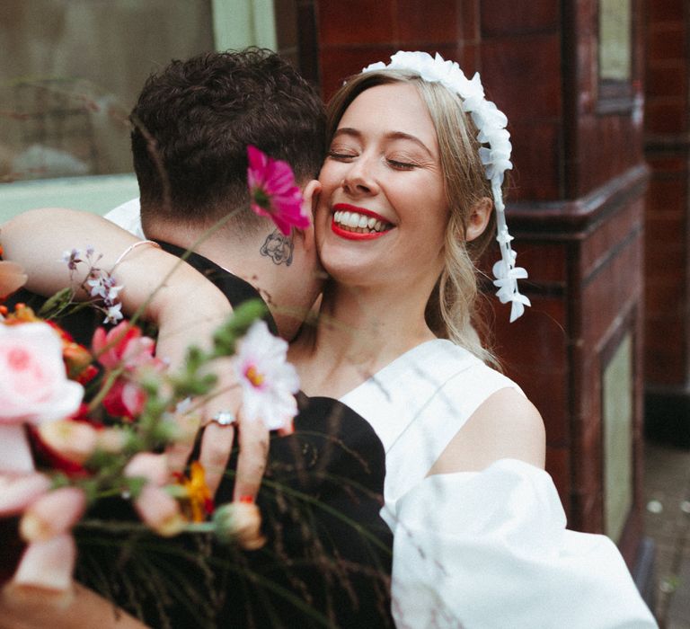 Bride wears red lipstick to match her bright and colourful bouquet as she hugs the groom