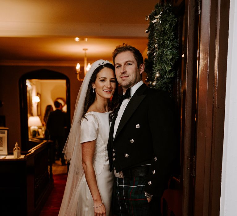 Bride and groom stand in the doorway of their wedding reception and look to camera whilst holding hands.