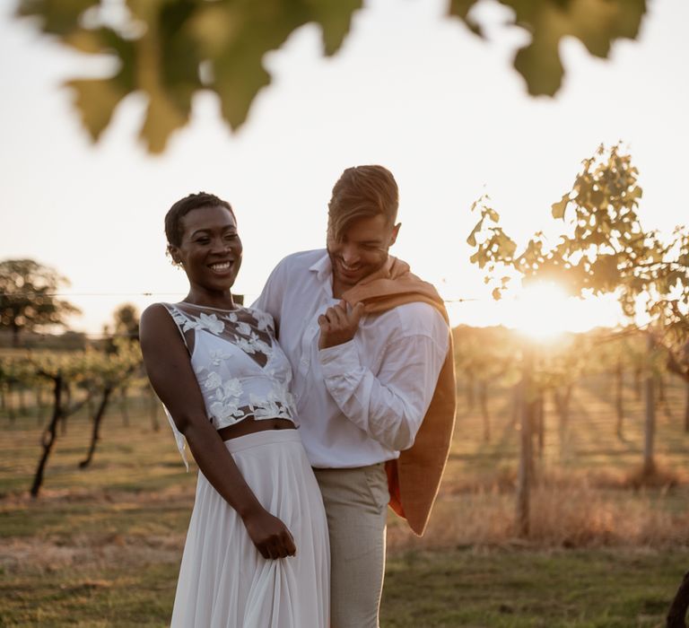 Gold hour portrait with black bride with short hair wearing bridal separates and groom in white shirt with jacket thrown over his shoulder 