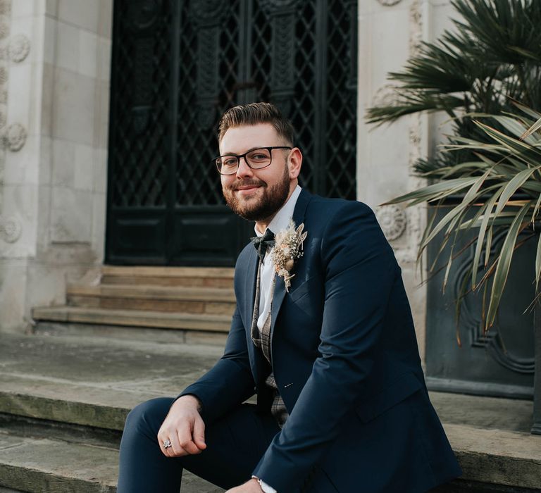 Smiling groom in navy suit with tartan waistcoat and dried flower buttonhole sits on steps outside