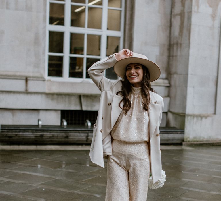 Brunette bride wears neutral coloured hat and sparkly wedding jumpsuit with blazer