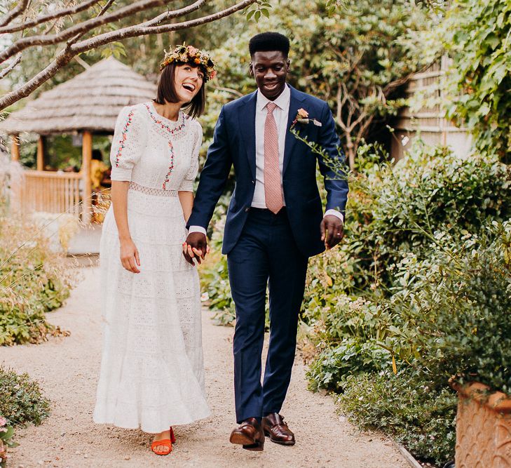 Groom in a navy suit and pink tie holding his brides hand in a white Broderie Anglaise wedding dress and colourful flower crown in a garden 