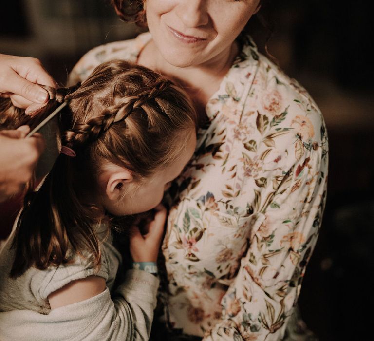 The bride in pyjamas getting ready and holding a small child having her hair braided.