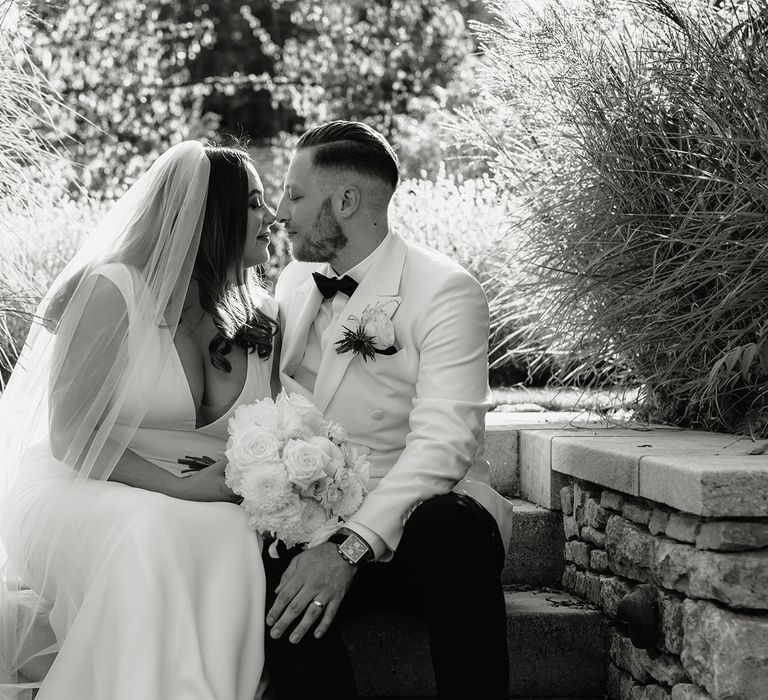 Bride in V neck Made With Love wedding dress and veil holding white rose bouquet sits outside with groom in white double breasted tuxedo jacket