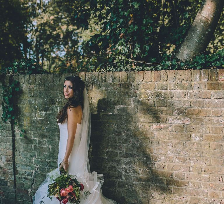 Bride stood against brick wall wearing white fishtail gown and pink and res rose bouquet 