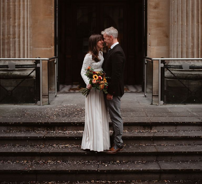 Bride in Self Portrait wedding dress holding colourful bouquet stands with groom on steps outside Bristol Registry Office