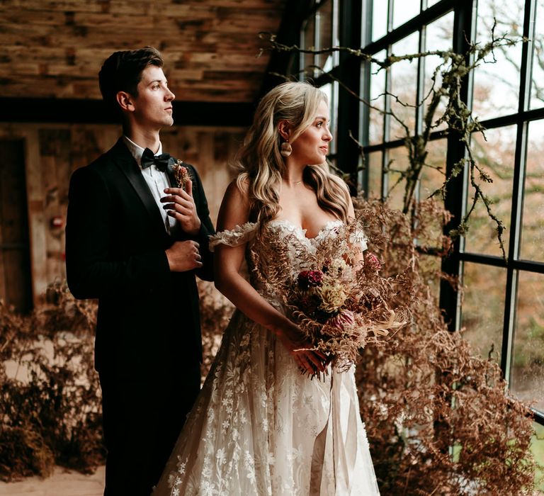 Groom in a black tuxedo standing at the window with his bride in an appliqué wedding dress surrounded by dried flower arrangements 