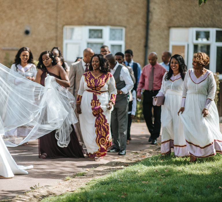 Bride in a princess wedding dress with long veil holding a white peony bouquet walking to the church with her bridal party 