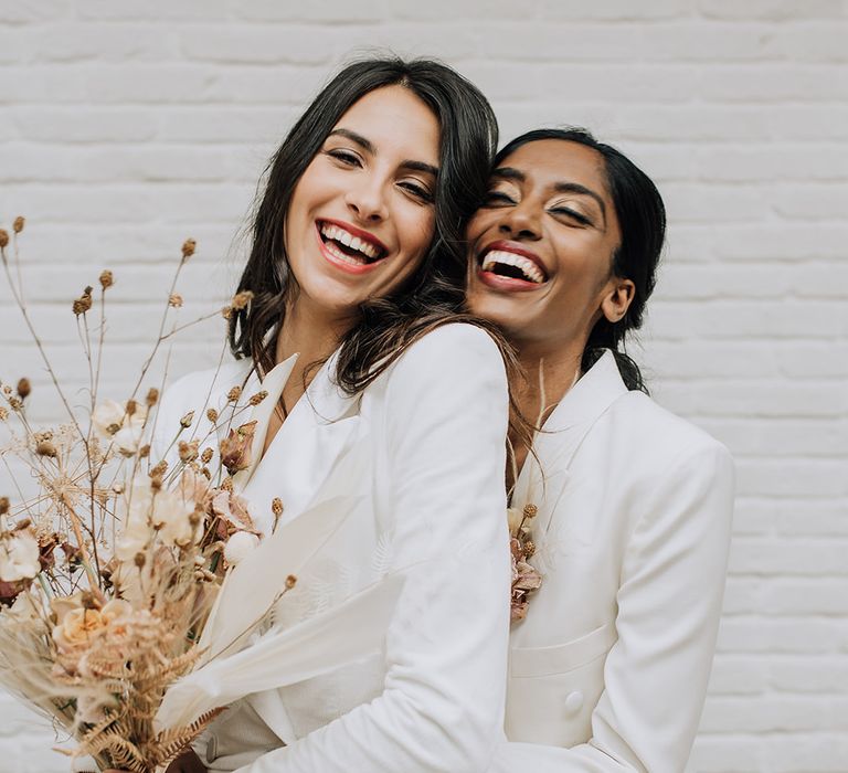 Two brides hugging and laughing at their all white wedding with dried flowers and bridal suits