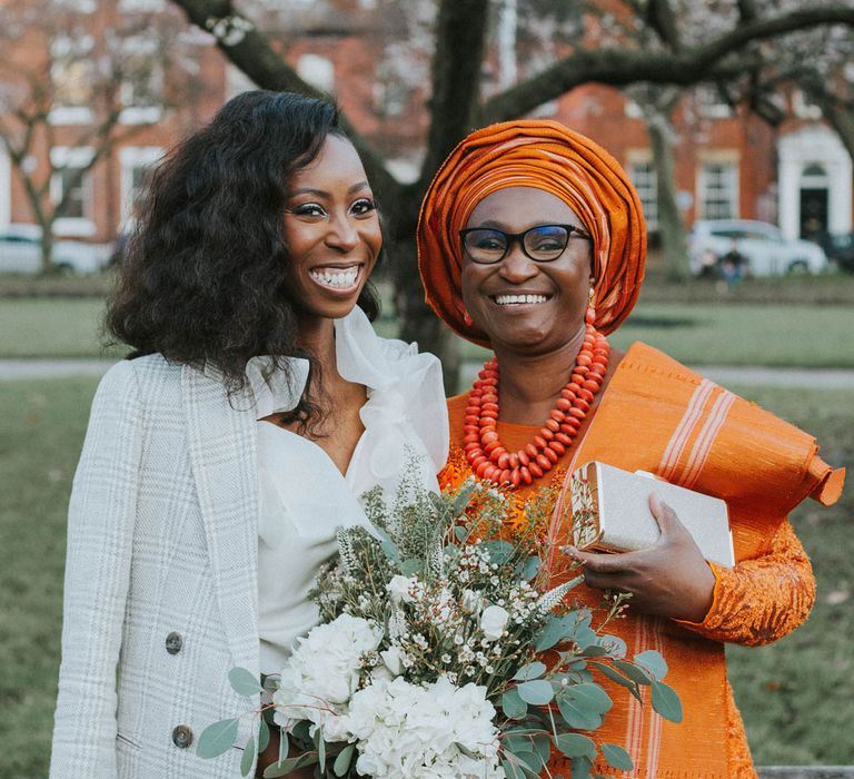 Bride with short curled hair in white Bec + Bride satin dress and grey checked blazer holds white and green wedding bouquet standing next to wedding guest in orange dress, beaded necklace and headscarf outside Bridge Community Church wedding