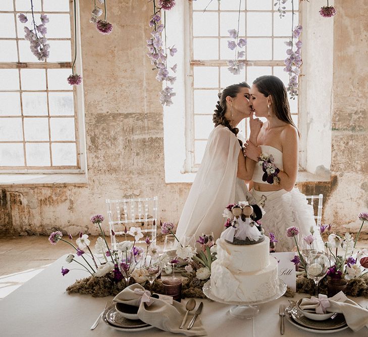 Two brides kissing at their Intimate sweetheart table with purple wedding flowers and iced wedding cake 