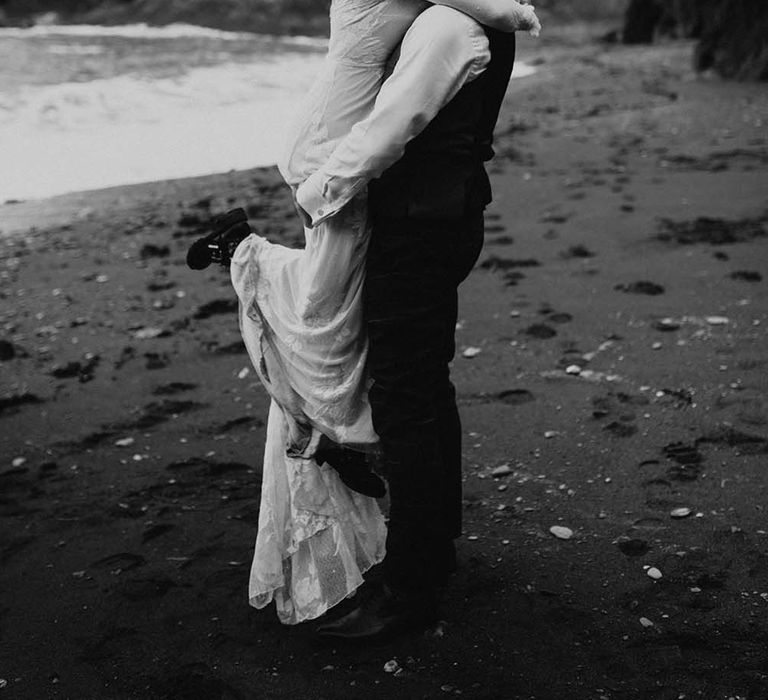 Black and white portrait of the groom picking up his bride on the beach in Devon