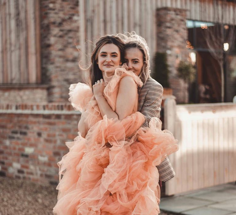 A lesbian couple hug for a wedding portrait. The woman in front wears a ruffled peach gown. Photography by Photography Events. 