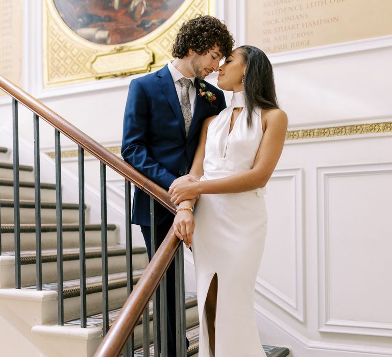 Bride & groom stand on staircase whilst kissing as bride wears high neck gown with split to the front