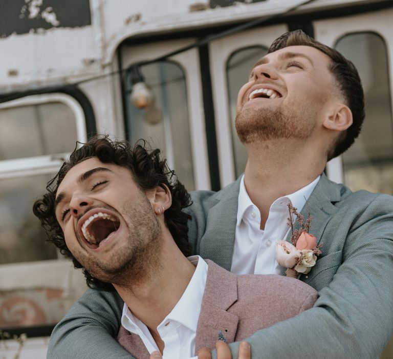 Grooms laugh with one another outdoors whilst wearing pastel suits