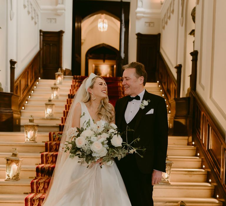 Bride looks lovingly at her father whilst smiling as they stand upon staircase before wedding ceremony