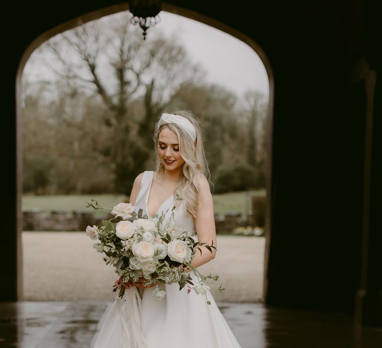 Blonde bride wears Suzanne Neville dress holding classical and elegant white bouquet with green foliage 