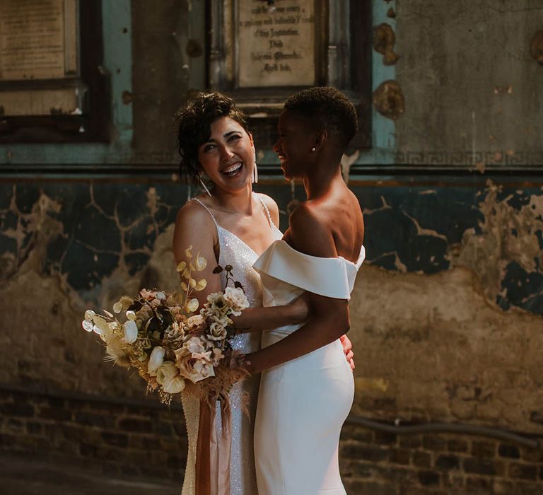 Two brides laughing and embracing holding a dried and fresh flower bouquet tied with ribbon 