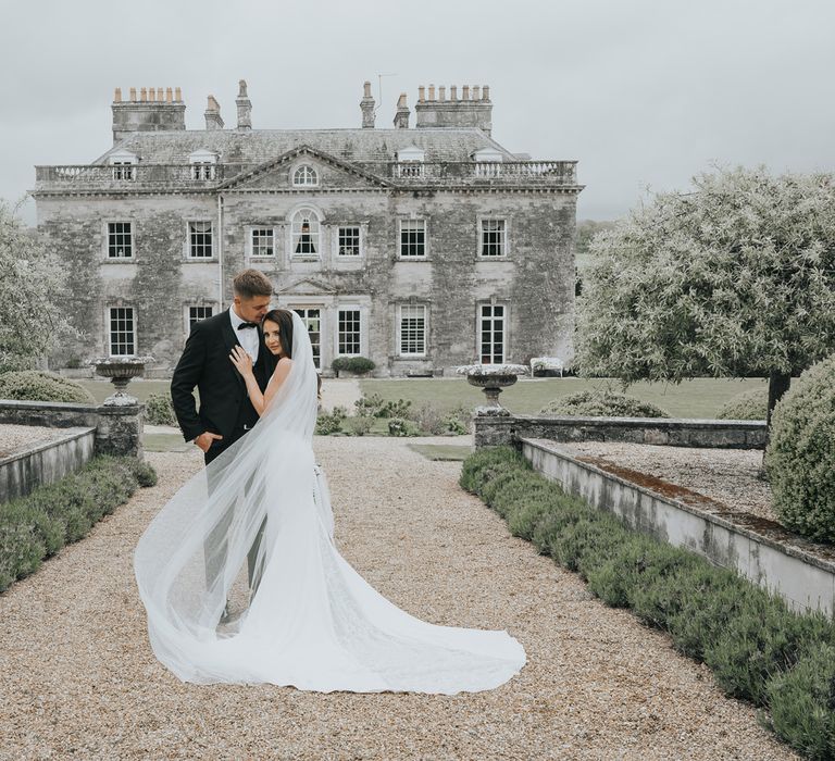 Bride in white Pronovias wedding dress with train and veil stands with hand on grooms chest as they stand on path in front of Came House Dorset after their wedding