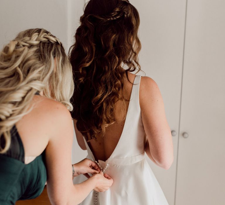 Bride with long brown curled hair with braid has her Elbeth Gillis low back wedding gown buttoned up by bridesmaid in green dress